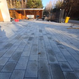 Artbruk - Układanie szarej kostki brukowej na posesji, widoczny pracownik w trakcie pracy, przyczepa z narzędziami, częściowo ukończona nawierzchnia.