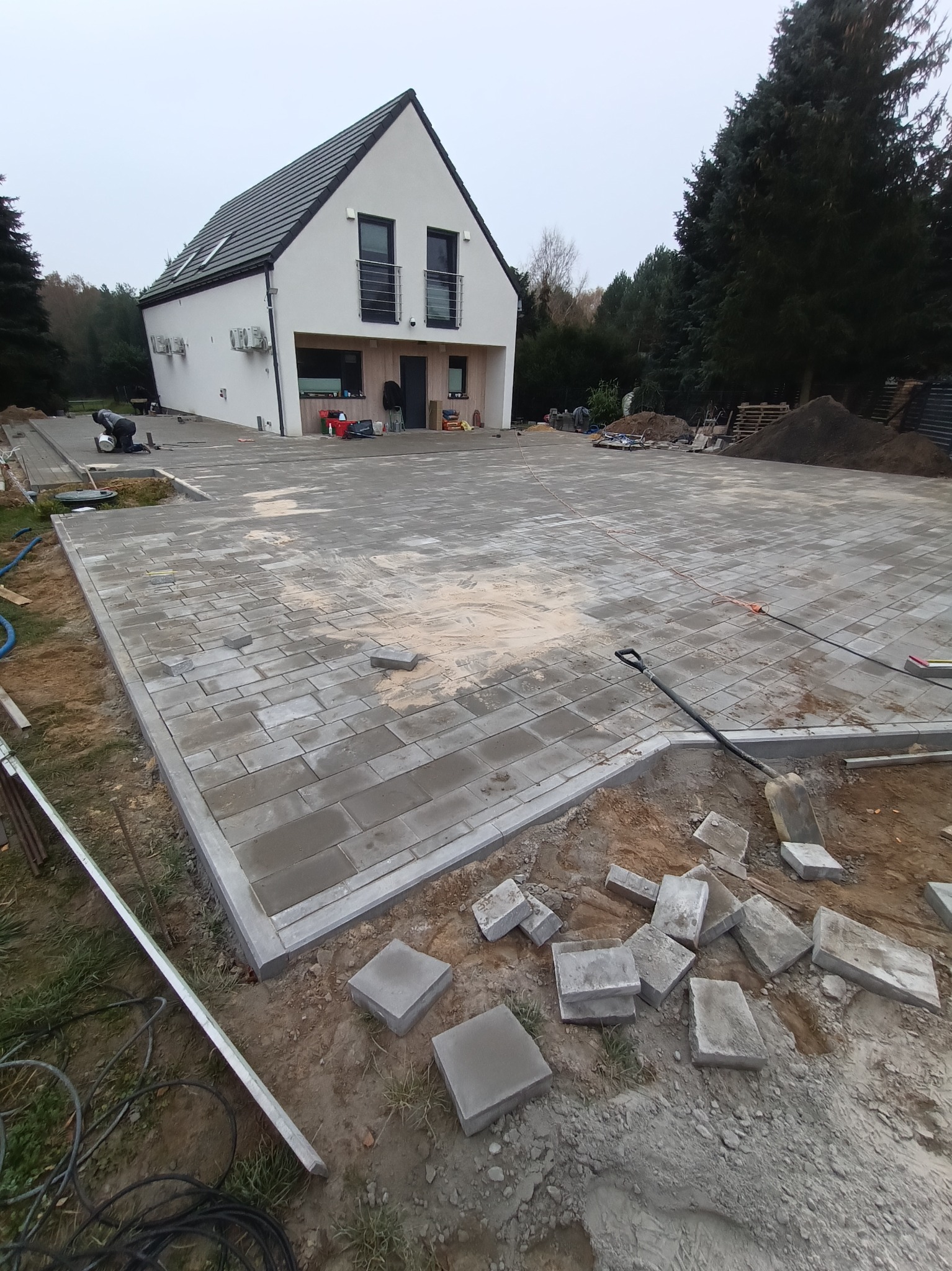 Rozległy podjazd z szarej kostki brukowej przed nowoczesnym, dwupiętrowym domem z białymi ścianami i czarnym dachem, częściowo wykończony, z widocznymi narzędziami i stertą kostek na pierwszym planie.