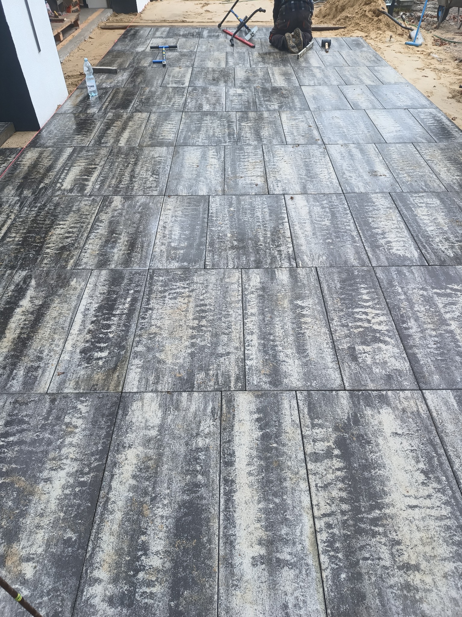 Ułożona nawierzchnia z szarych, prostokątnych płyt brukowych o nieregularnym wzorze, widoczne narzędzia pracy i pracownik w tle, butelka wody obok ściany budynku.
