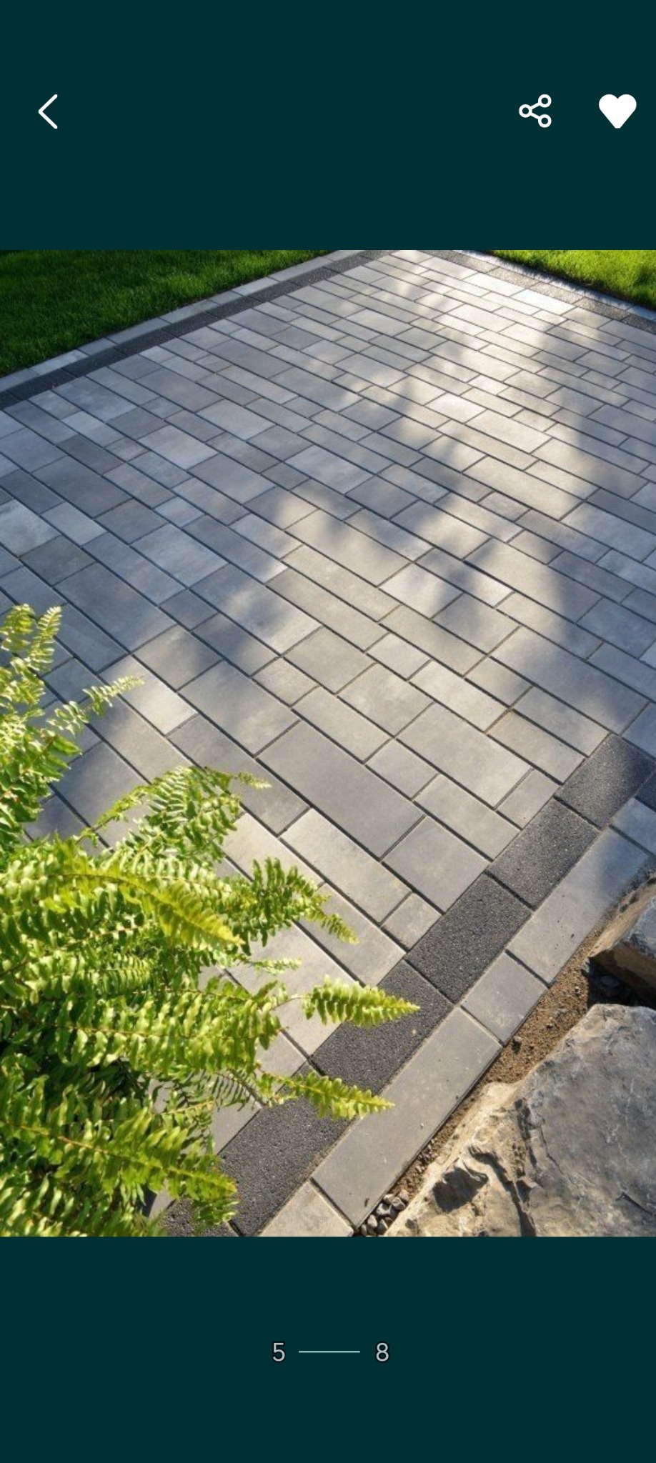 Świeżo ułożona, szara kostka brukowa w kształcie prostokątów, obramowana ciemniejszym pasem, z zieloną paprocią w rogu i fragmentem kamienia w tle.