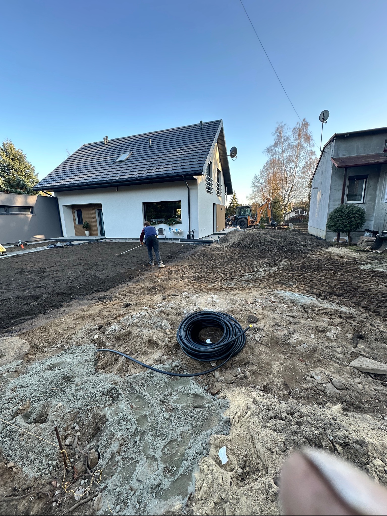 Przygotowanie terenu pod trawnik przed nowoczesnym domem: mężczyzna wyrównuje ziemię grabiami, w tle widoczny sprzęt budowlany i zwój węża.