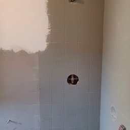 Tomasz Kruk Kompleksowe wykończenia wnętrz - Ściana łazienki w trakcie malowania i kładzenia płytek; widoczne fragmenty surowej ściany, pomalowanej częściowo na biało, oraz fragmentu z szarymi płytkami, z otworem na rurę i zawór; na podłodze...