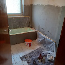 Tomasz Kruk Kompleksowe wykończenia wnętrz - Łazienka w trakcie prac przygotowawczych do wykończenia: zabezpieczona folią wanna, wiadro farby Alpina, narzędzia na podłodze, częściowo wyszpachlowane ściany, widoczne wyprowadzenia instalacji.