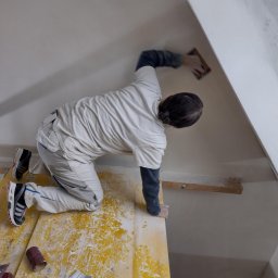 Tomasz Kruk Kompleksowe wykończenia wnętrz - Pracownik na rusztowaniu szlifuje skośną ścianę poddasza, używając pacy z papierem ściernym; widoczne narzędzia i resztki materiałów na żółtym rusztowaniu.