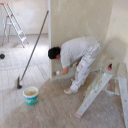 Tomasz Kruk Kompleksowe wykończenia wnętrz - Pracownik w białym kombinezonie, pochylony, nakłada masę na ścianę w rogu pomieszczenia; obok drabina, wiadro z masą i narzędzie do rozprowadzania masy na podłodze.