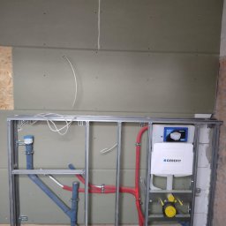 Tomasz Kruk Kompleksowe wykończenia wnętrz - Stelaż podtynkowy Geberit zamontowany na tle surowej ściany z płyt gipsowo-kartonowych, widoczne rury doprowadzające i odprowadzające wodę, instalacja w trakcie montażu.