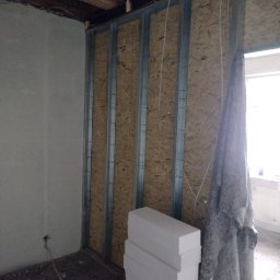 Tomasz Kruk Kompleksowe wykończenia wnętrz - Konstrukcja ścianki działowej z metalowych profili i płyty OSB, widoczne przewody elektryczne, stos białego styropianu przy ścianie, fragment okna z zasłoniętą szybą.