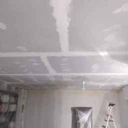 Pokój w trakcie prac wykończeniowych: sufit z widocznymi łączeniami płyt gipsowo-kartonowych i wstępnie nałożoną warstwą masy szpachlowej, zabezpieczone folią meble, drabina w rogu pomieszczenia.