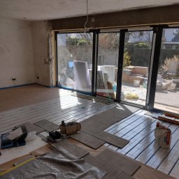 Tomasz Kruk Kompleksowe wykończenia wnętrz - Wnętrze w trakcie prac wykończeniowych: układanie płytek podłogowych na ogrzewaniu podłogowym; widoczne narzędzia, materiały i otwarte przesuwne drzwi tarasowe z widokiem na ogród.