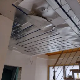 Montaż folii aluminiowej na suficie podwieszanym, widoczne profile i fragment instalacji elektrycznej, w tle schody i okno.