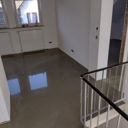 Tomasz Kruk Kompleksowe wykończenia wnętrz - Wylany samopoziomujący podkład podłogowy w jasnym pomieszczeniu z oknami, widoczne grzejniki i fragment schodów z metalową balustradą.