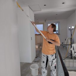 Tomasz Kruk Kompleksowe wykończenia wnętrz - Kobieta maluje ścianę wałkiem na długim wysięgniku w jasnym wnętrzu, widoczne puszki z farbą i lampa oświetleniowa, malarskie spodnie z plamami farby.