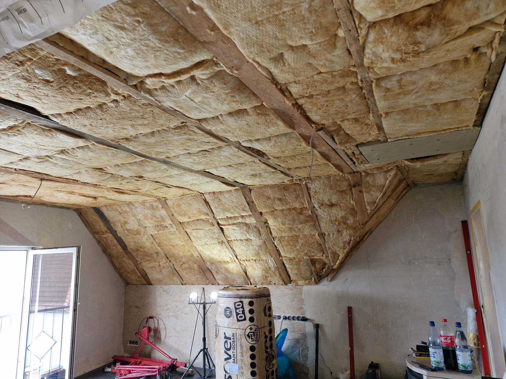 Poddasze w trakcie ocieplania wełną mineralną, widoczne drewniane belki konstrukcyjne, rolka wełny Isover, narzędzia i poziomice budowlane w tle.