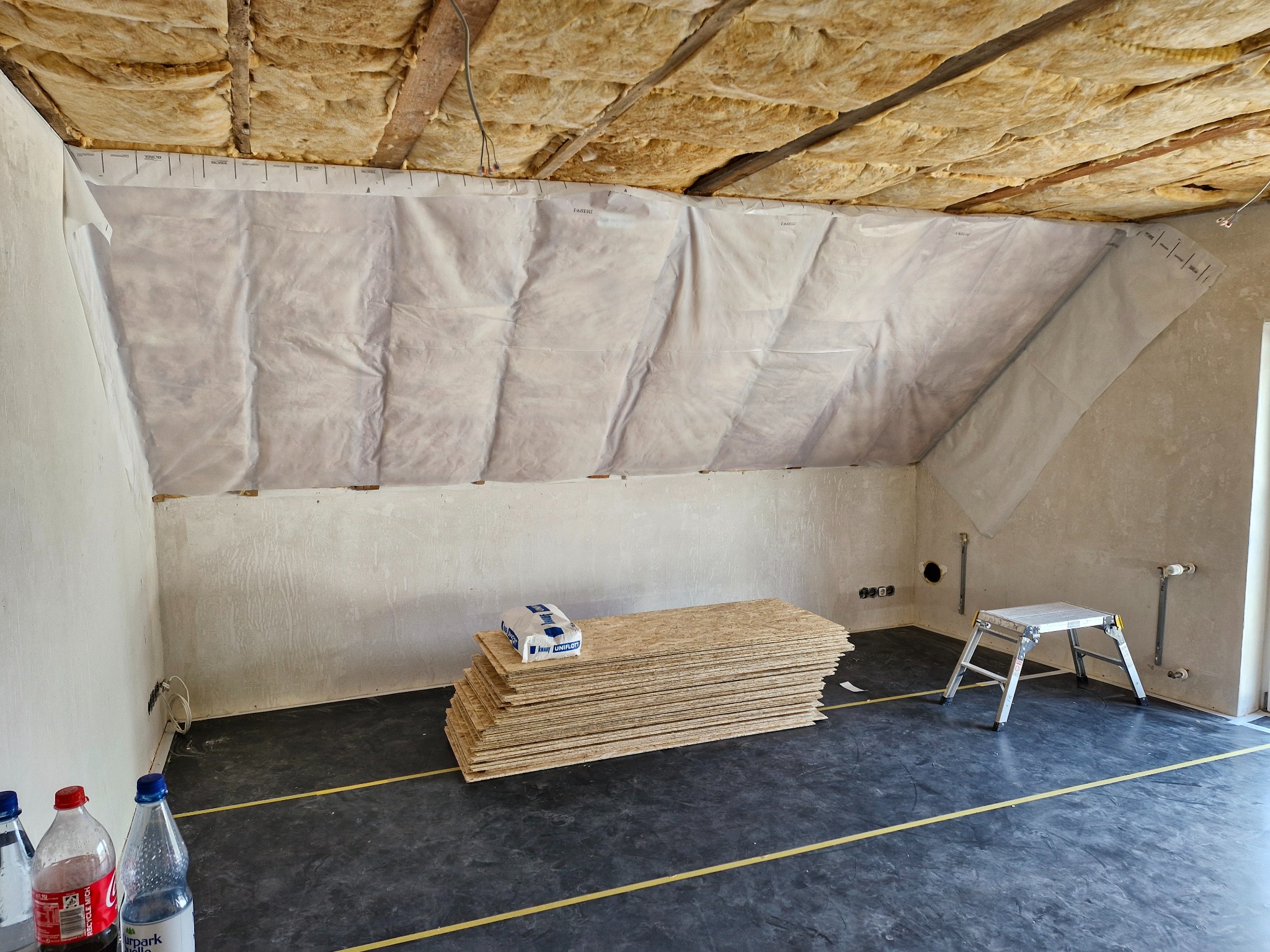 Poddasze w trakcie ocieplania: widoczna wełna mineralna pod dachem, folia paroizolacyjna na ścianach, stos płyt OSB z opakowaniem zaprawy, oraz aluminiowy podest.