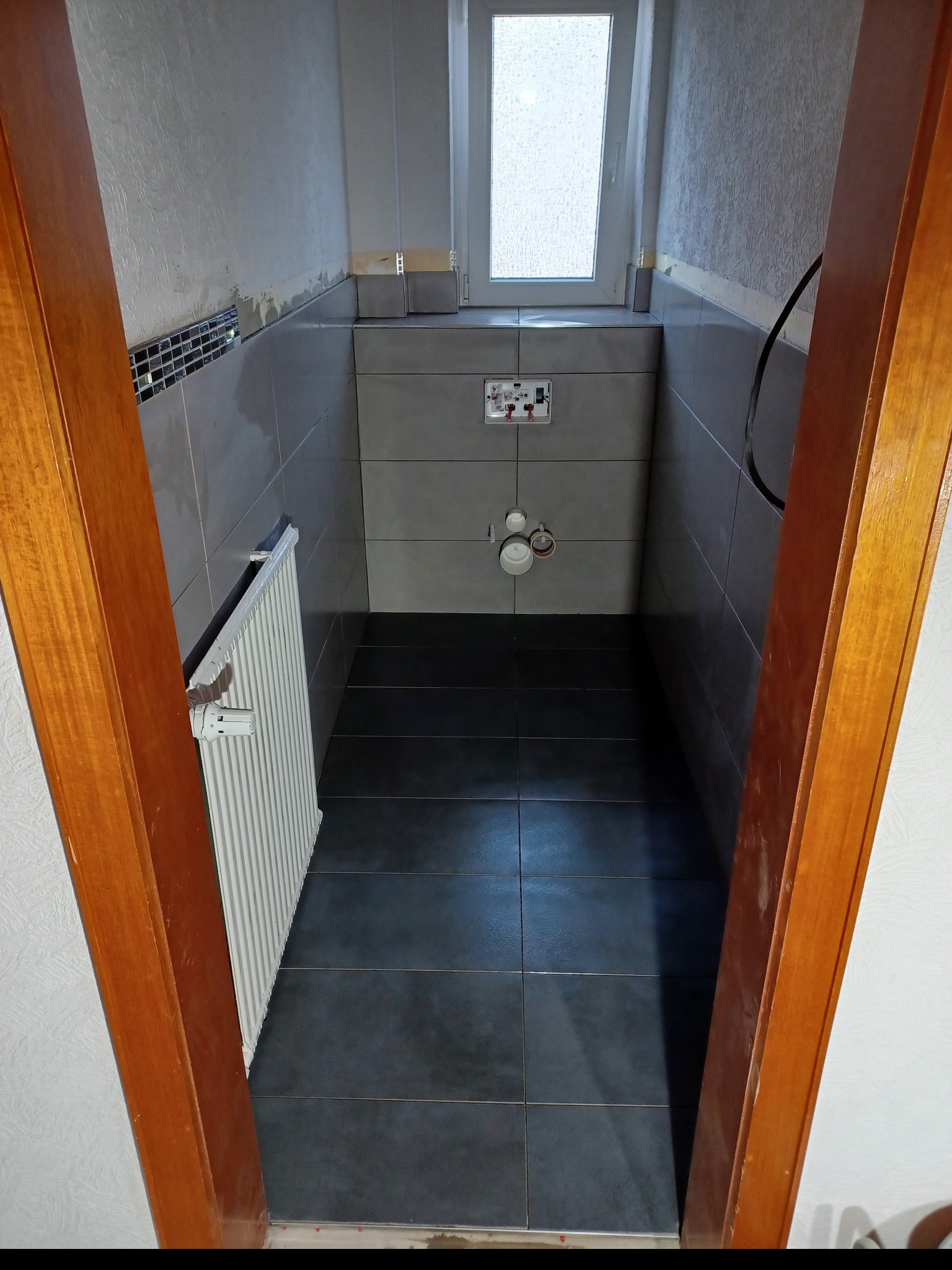 Wąska łazienka w trakcie remontu, widoczne szare płytki na ścianach i podłodze, zamontowany grzejnik i okno, częściowo położona mozaika, instalacja pod umywalkę i toaletę.