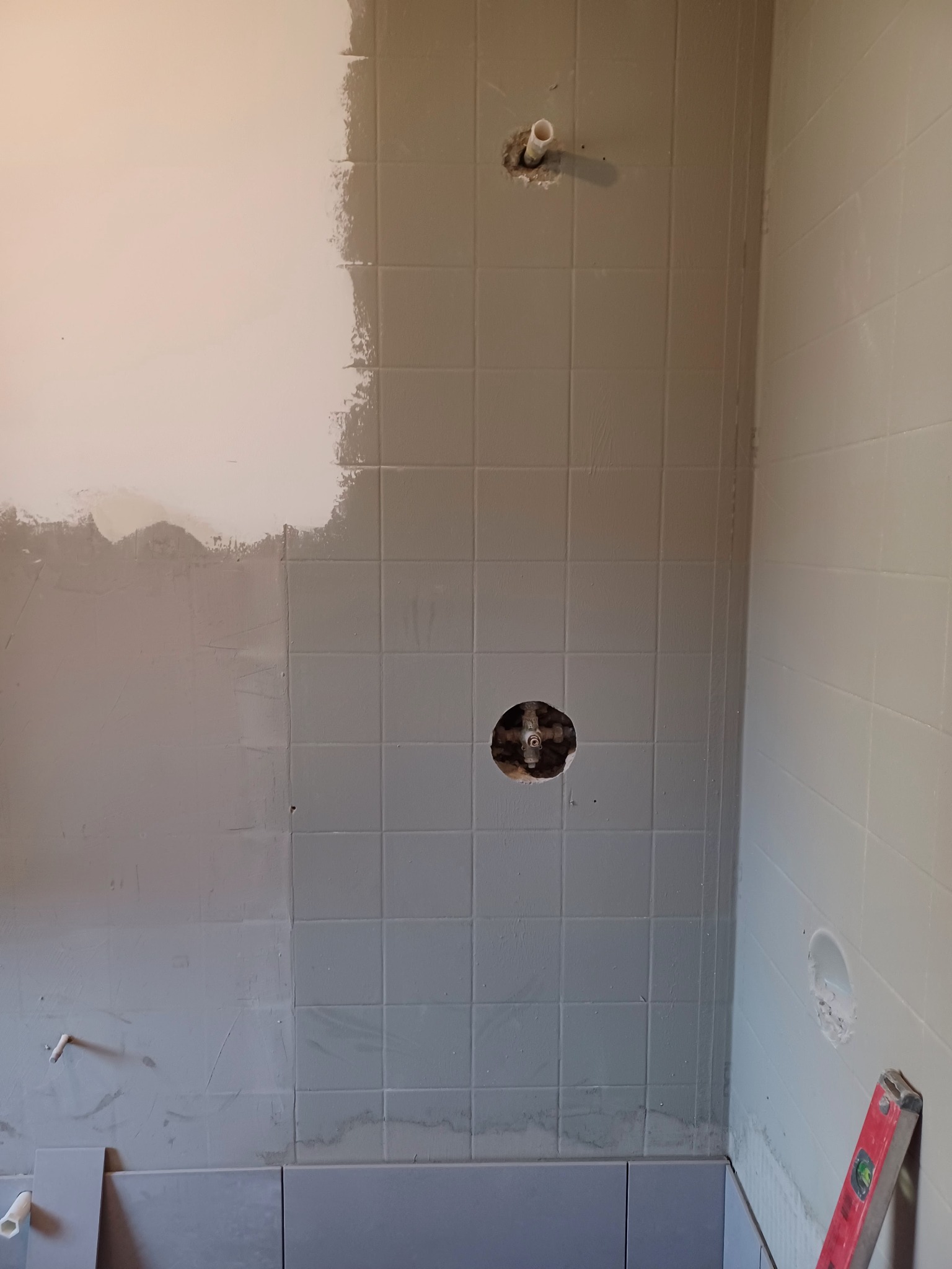 Ściana łazienki w trakcie malowania i kładzenia płytek; widoczne fragmenty surowej ściany, pomalowanej częściowo na biało, oraz fragmentu z szarymi płytkami, z otworem na rurę i zawór; na podłodze...