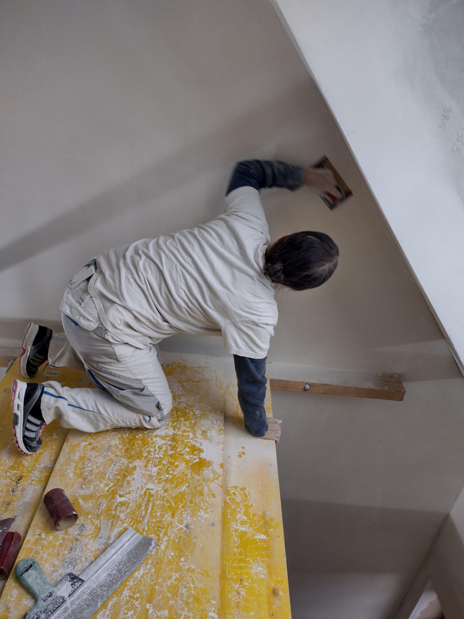 Pracownik na rusztowaniu szlifuje skośną ścianę poddasza, używając pacy z papierem ściernym; widoczne narzędzia i resztki materiałów na żółtym rusztowaniu.