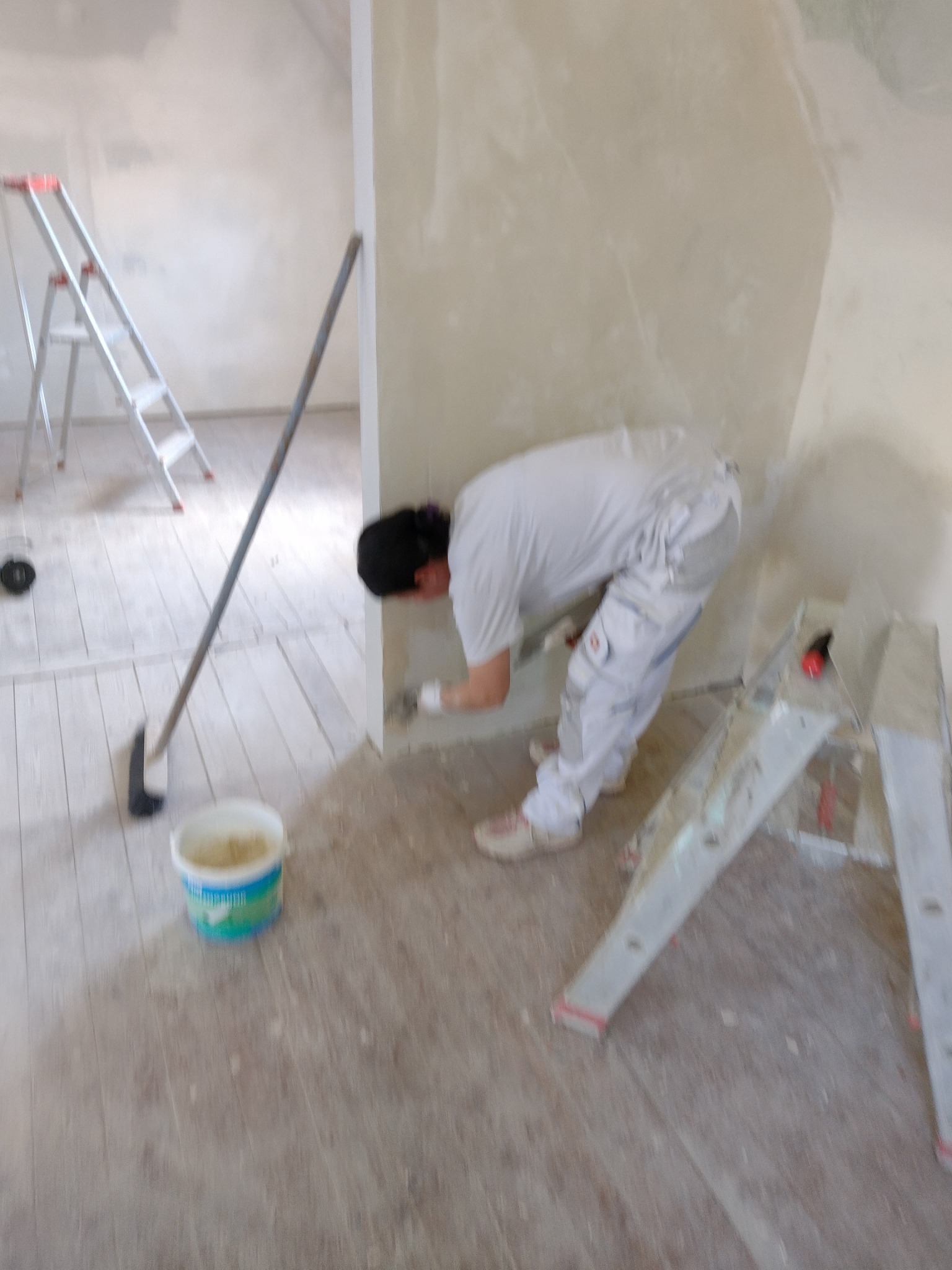 Pracownik w białym kombinezonie, pochylony, nakłada masę na ścianę w rogu pomieszczenia; obok drabina, wiadro z masą i narzędzie do rozprowadzania masy na podłodze.