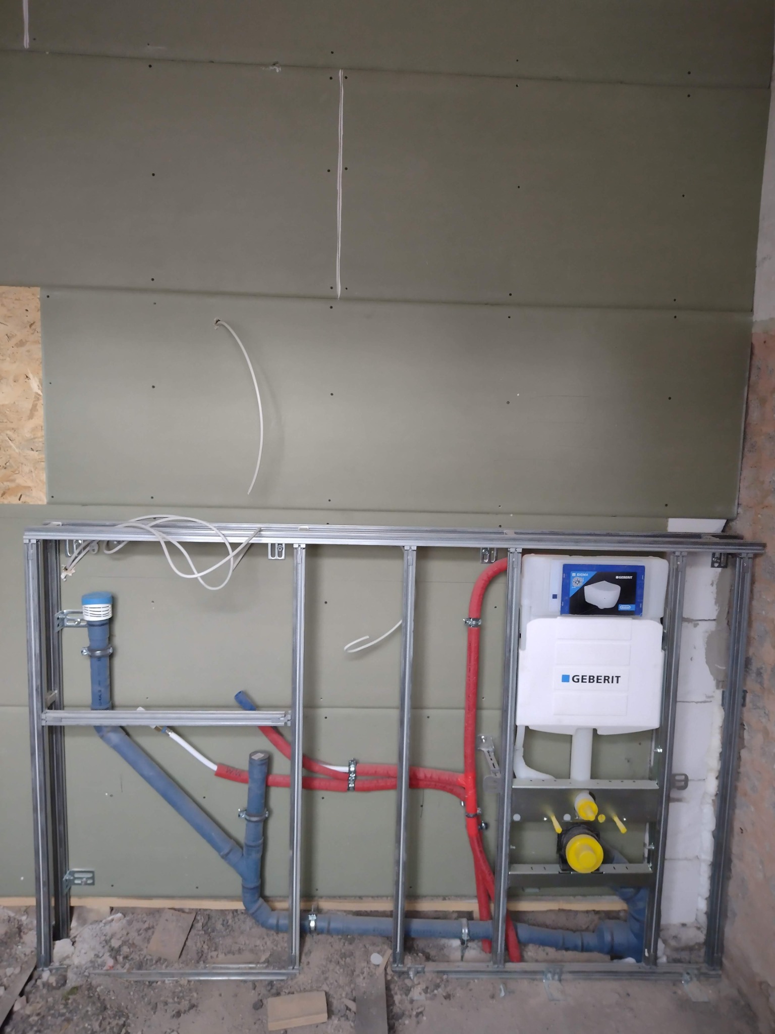 Stelaż podtynkowy Geberit zamontowany na tle surowej ściany z płyt gipsowo-kartonowych, widoczne rury doprowadzające i odprowadzające wodę, instalacja w trakcie montażu.