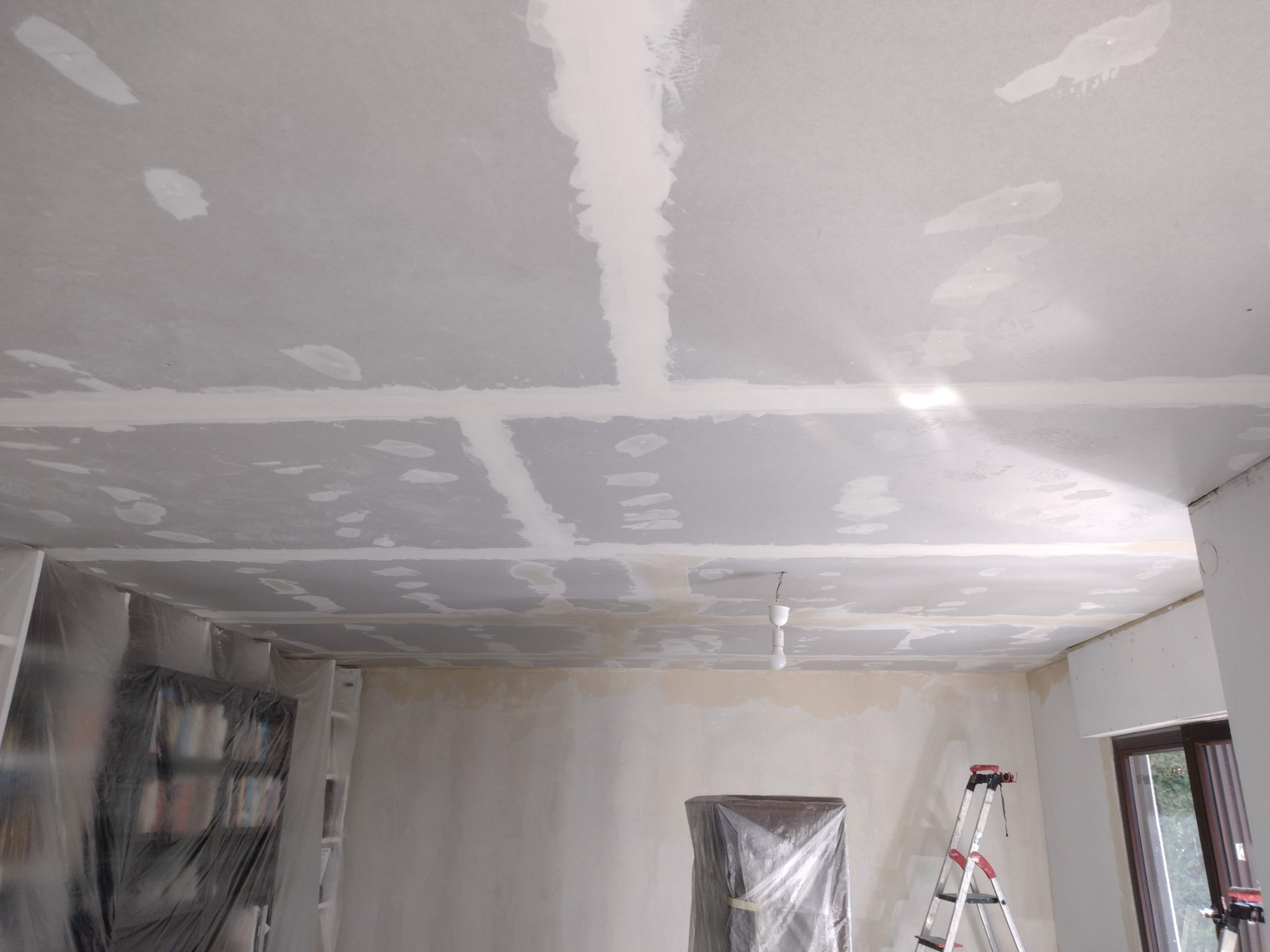 Pokój w trakcie prac wykończeniowych: sufit z widocznymi łączeniami płyt gipsowo-kartonowych i wstępnie nałożoną warstwą masy szpachlowej, zabezpieczone folią meble, drabina w rogu pomieszczenia.