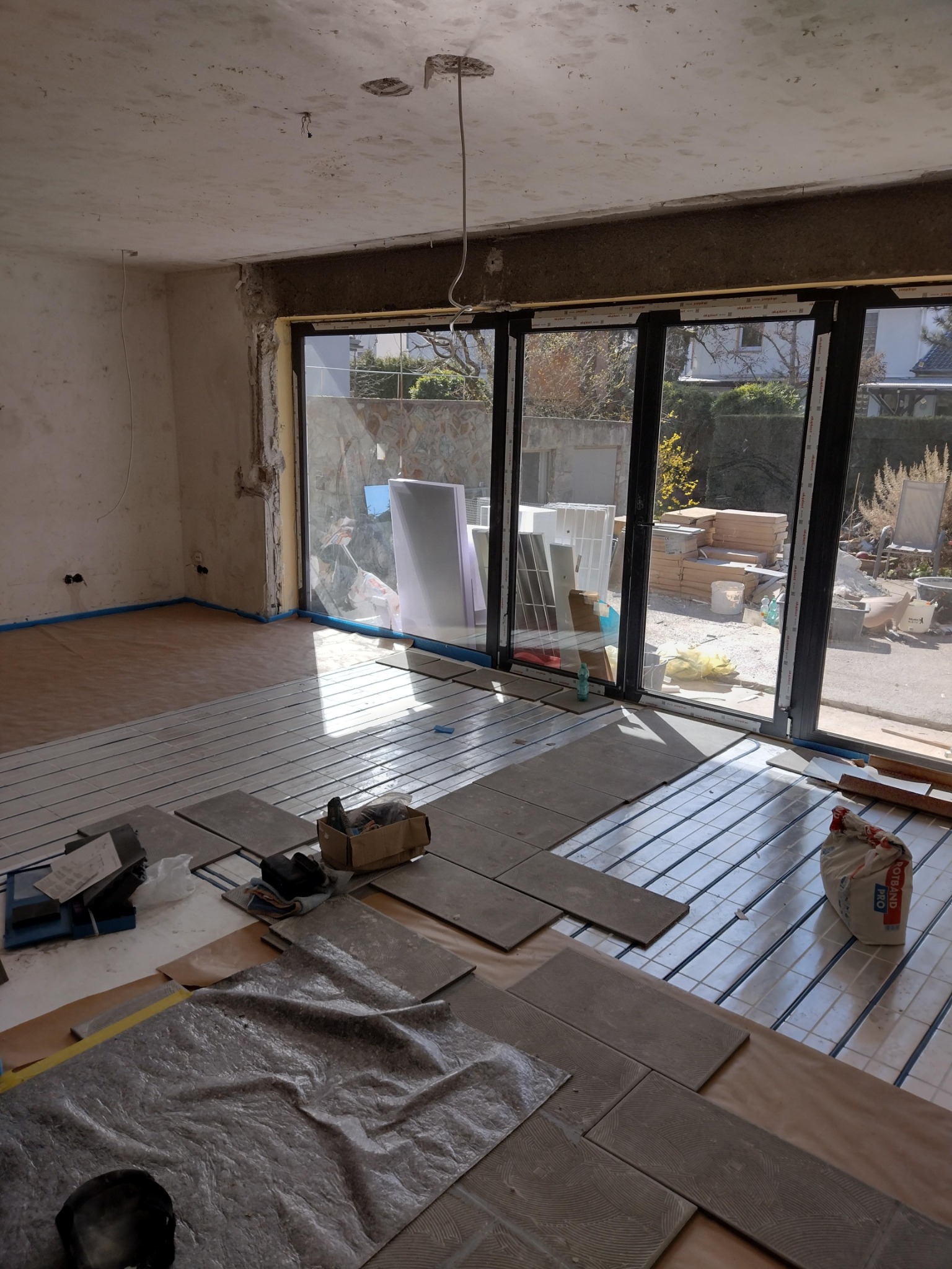 Wnętrze w trakcie prac wykończeniowych: układanie płytek podłogowych na ogrzewaniu podłogowym; widoczne narzędzia, materiały i otwarte przesuwne drzwi tarasowe z widokiem na ogród.