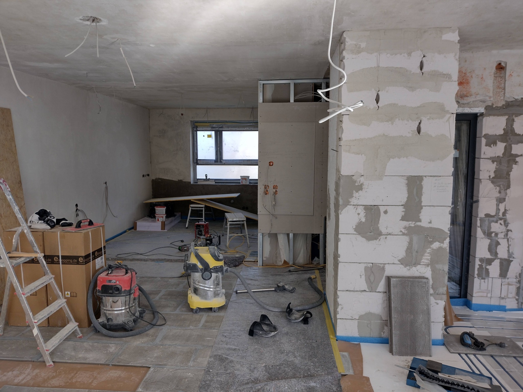 Wnętrze w trakcie prac wykończeniowych: widoczne surowe ściany z betonu komórkowego, przygotowane do dalszych prac, z instalacjami elektrycznymi, częściowo położona podłoga, drabina, dwa odkurzacze...