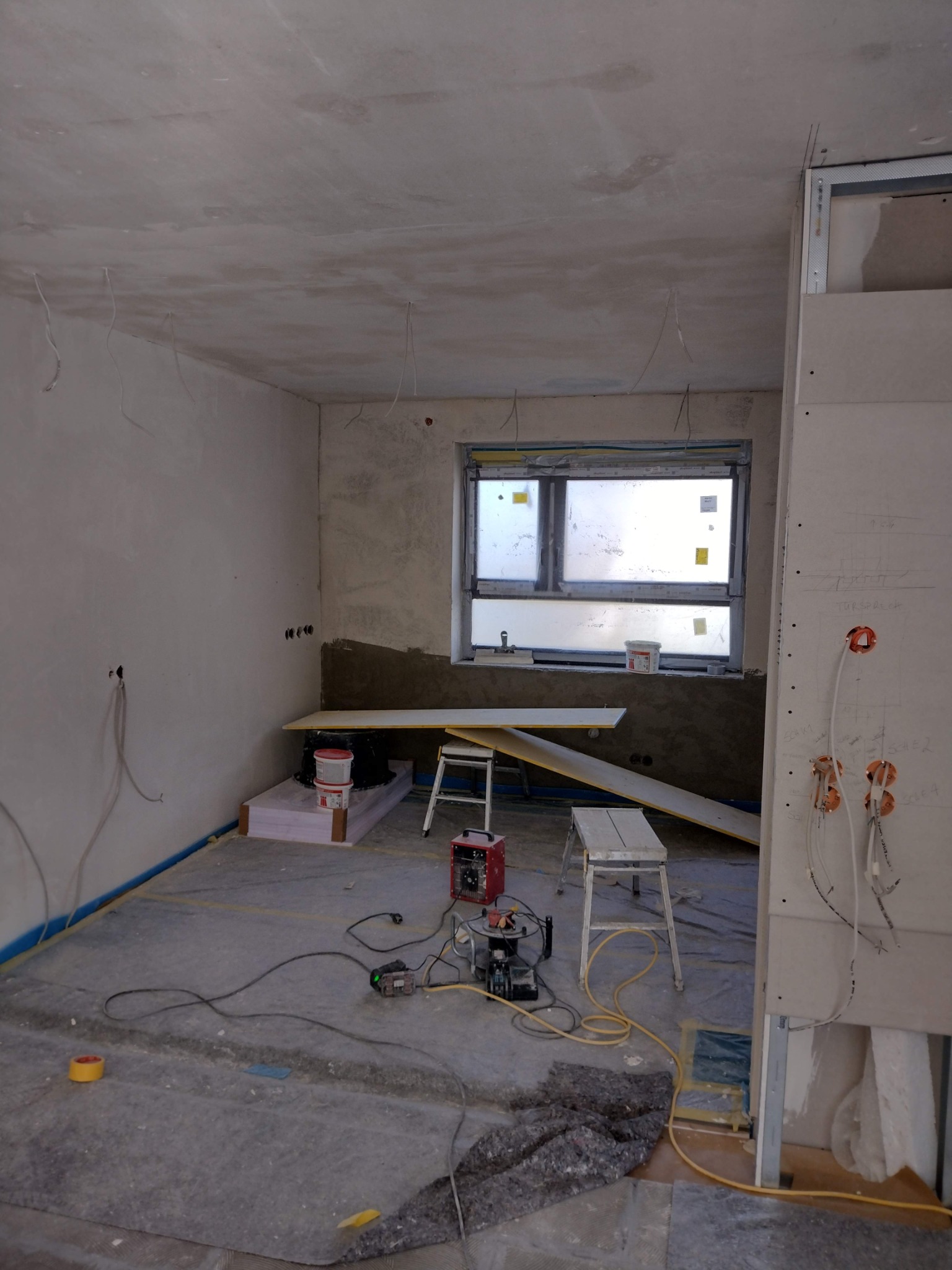 Wnętrze w trakcie prac wykończeniowych: ściany po szpachlowaniu, przygotowane do malowania, widoczne wystające kable elektryczne, okno z naklejonymi foliami, narzędzia i materiały budowlane...