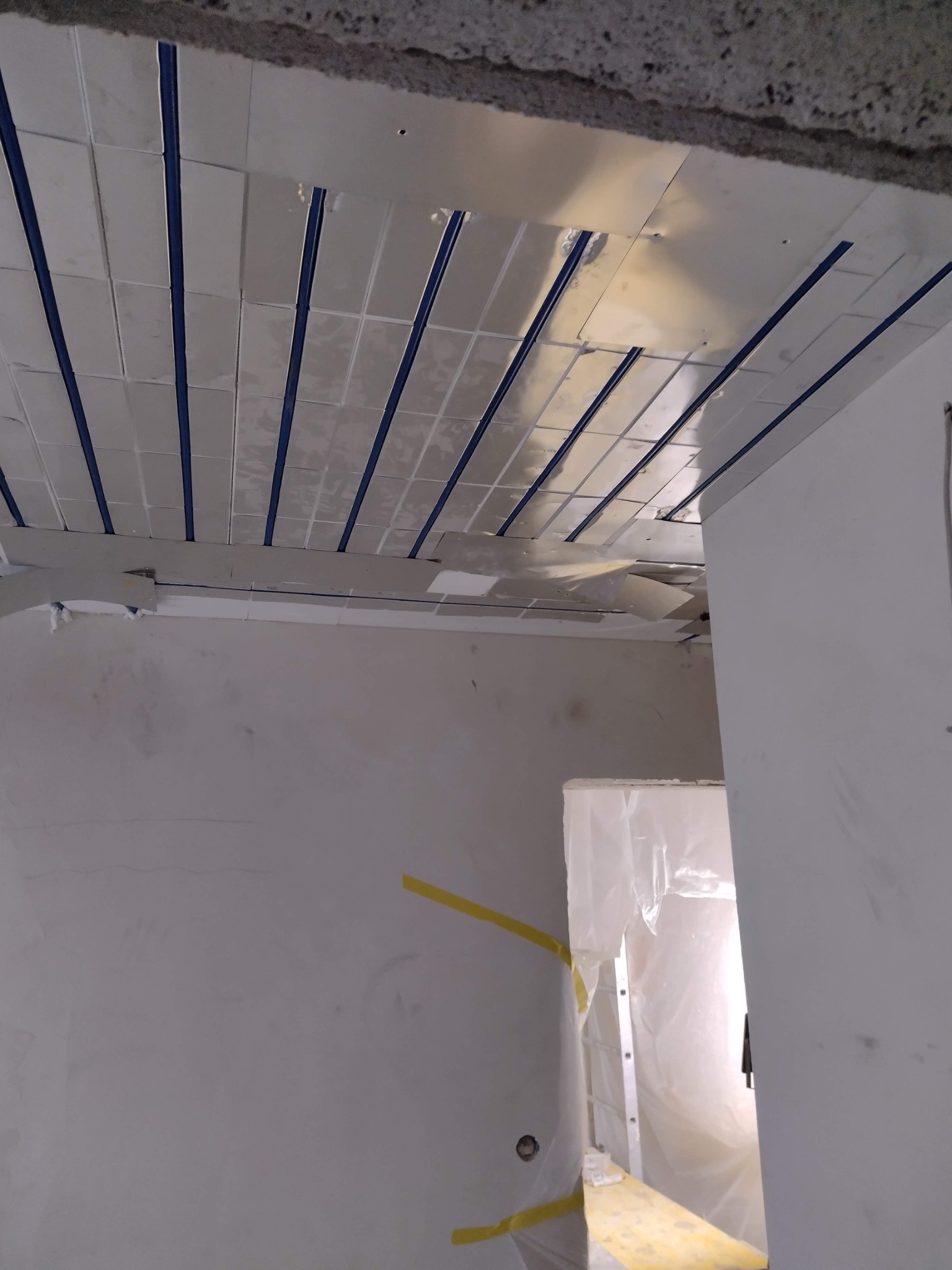 Montaż sufitu napinanego z białych paneli oddzielonych niebieskimi listwami, widoczne niedociągnięcia i elementy konstrukcyjne, ściany przygotowane do dalszych prac wykończeniowych.