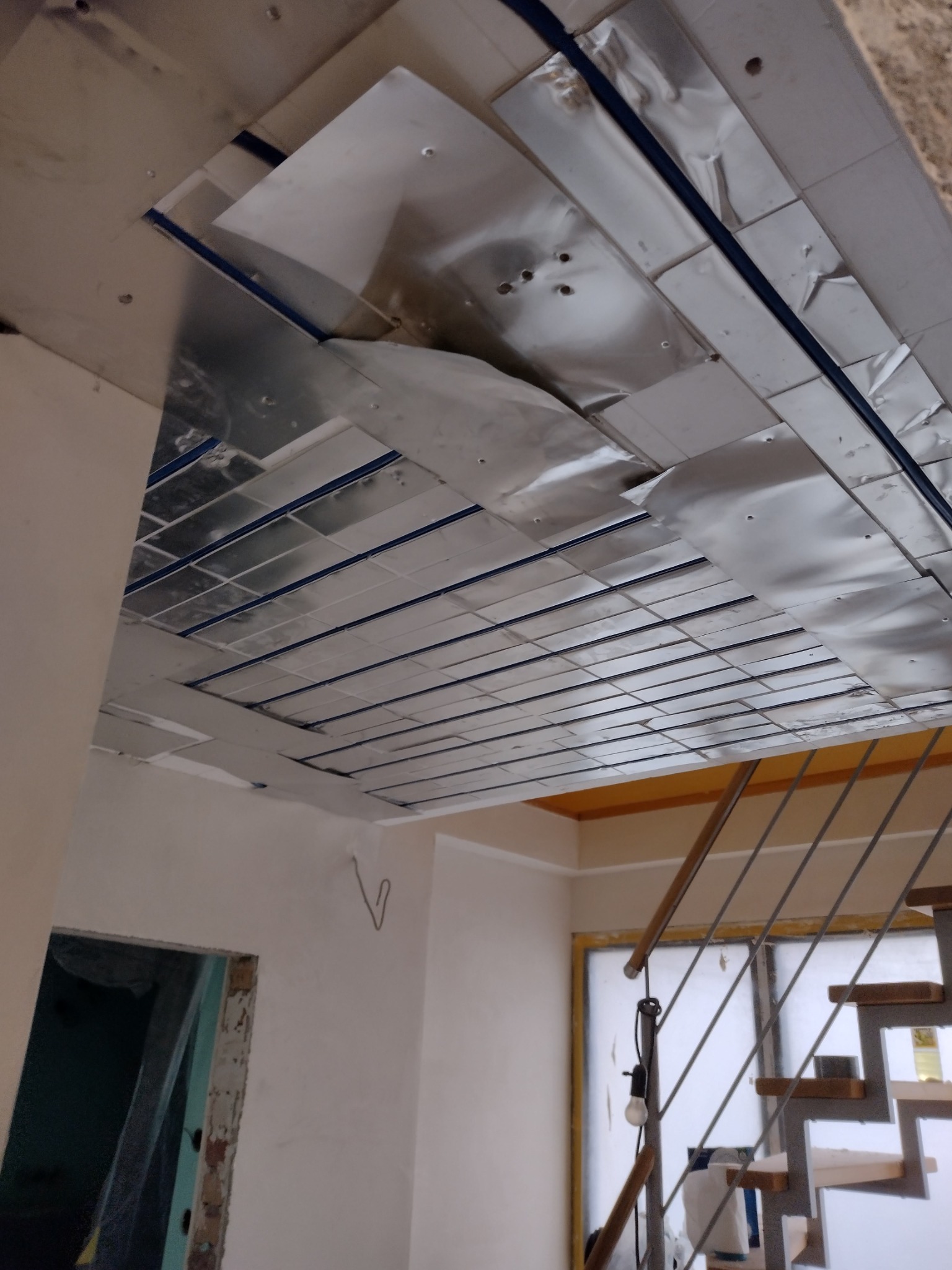 Montaż folii aluminiowej na suficie podwieszanym, widoczne profile i fragment instalacji elektrycznej, w tle schody i okno.
