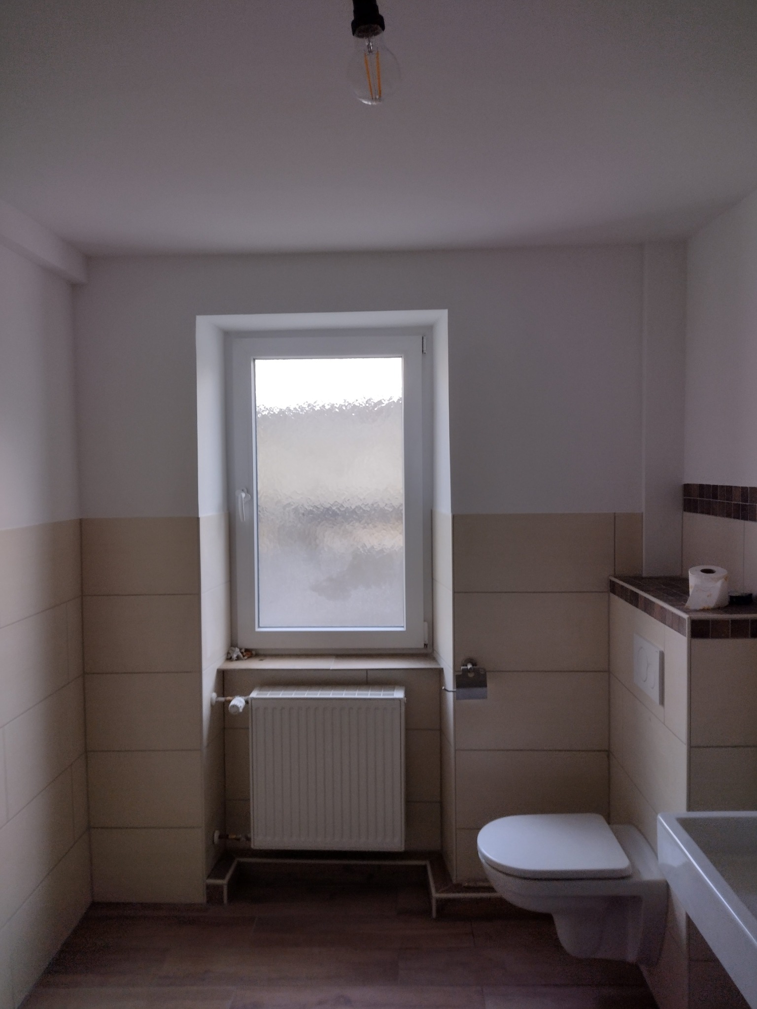 Wykończona łazienka w Lubawce z beżowymi płytkami, białym oknem z matową szybą, grzejnikiem panelowym i podwieszaną toaletą, widok na sufit z wiszącą żarówką.