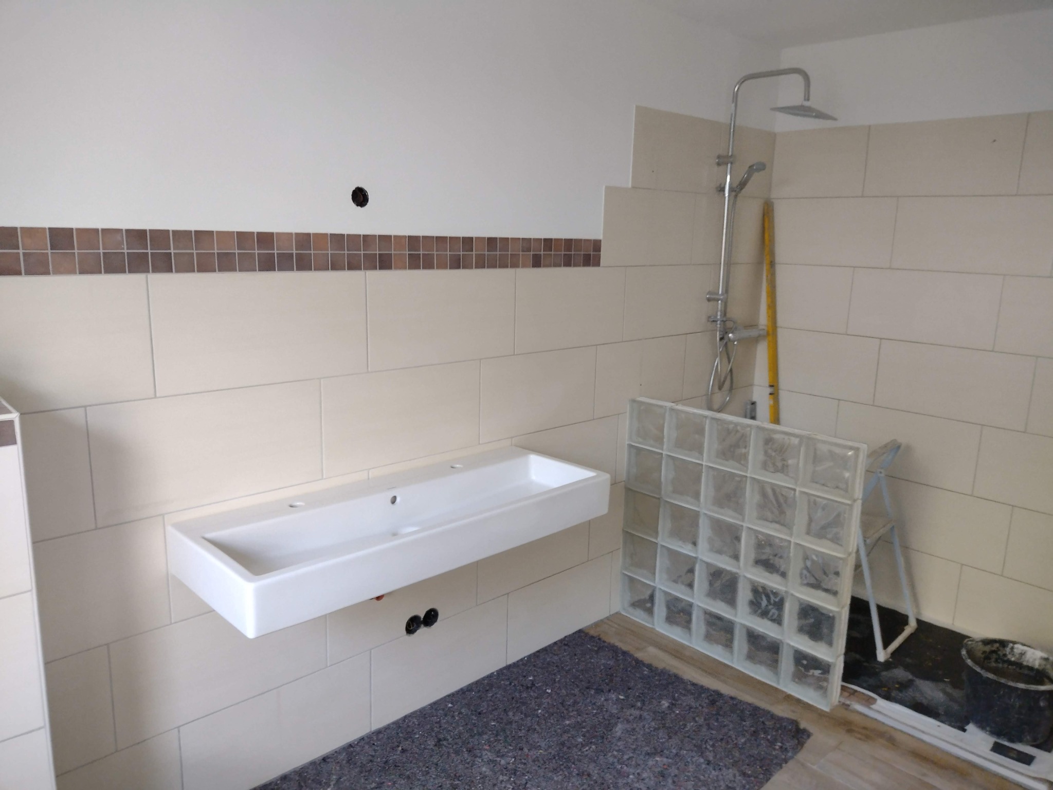 Montaż umywalki wiszącej w łazience, widoczna mozaika dekoracyjna, częściowo ułożone płytki, szklane luksfery i armatura prysznicowa w trakcie instalacji.