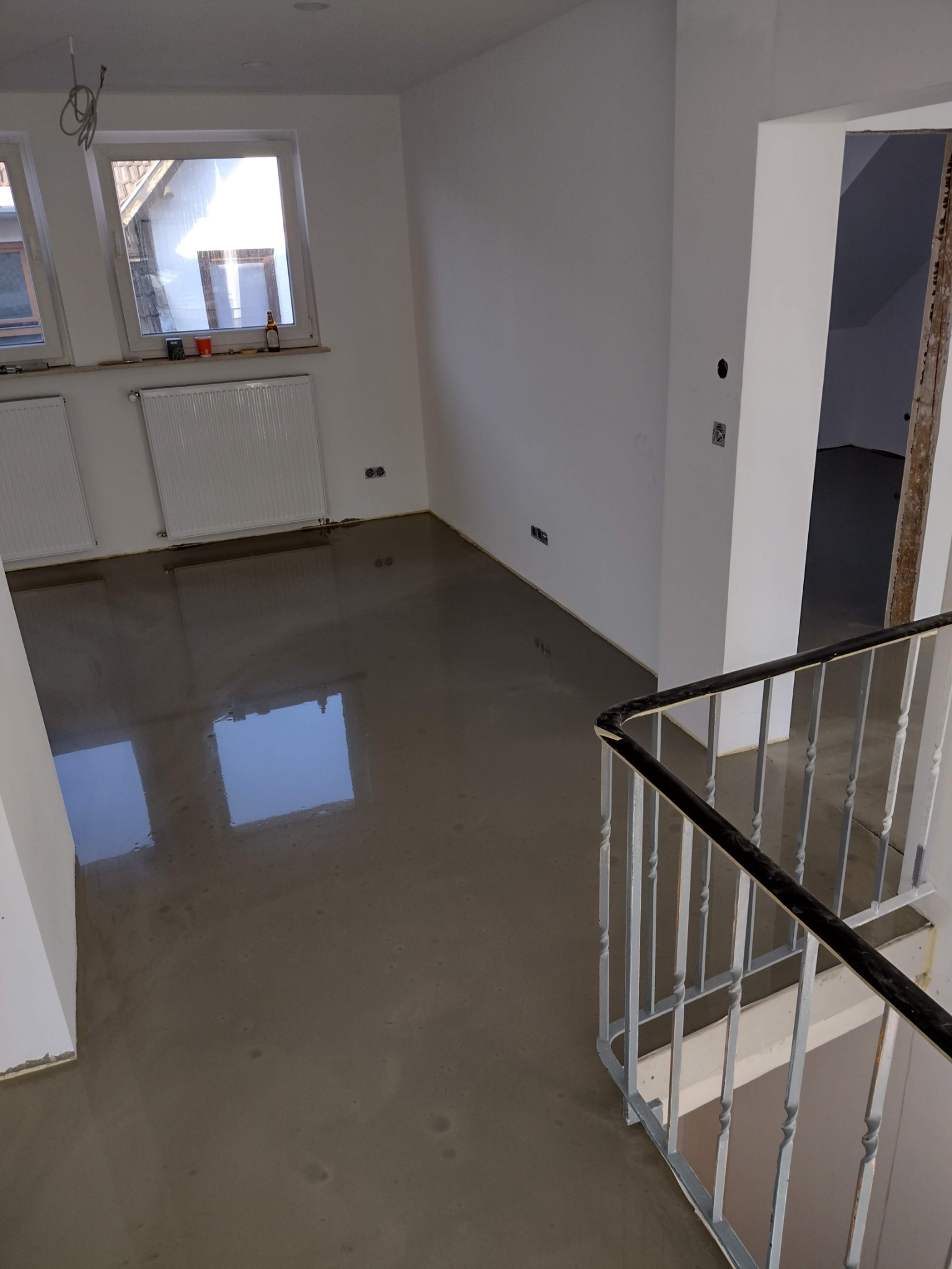 Wylany samopoziomujący podkład podłogowy w jasnym pomieszczeniu z oknami, widoczne grzejniki i fragment schodów z metalową balustradą.