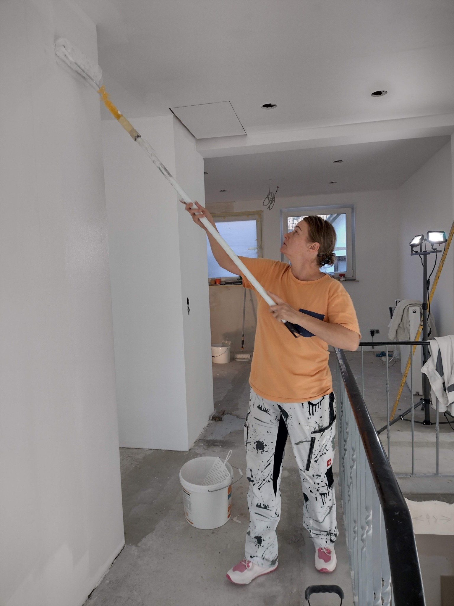 Kobieta maluje ścianę wałkiem na długim wysięgniku w jasnym wnętrzu, widoczne puszki z farbą i lampa oświetleniowa, malarskie spodnie z plamami farby.