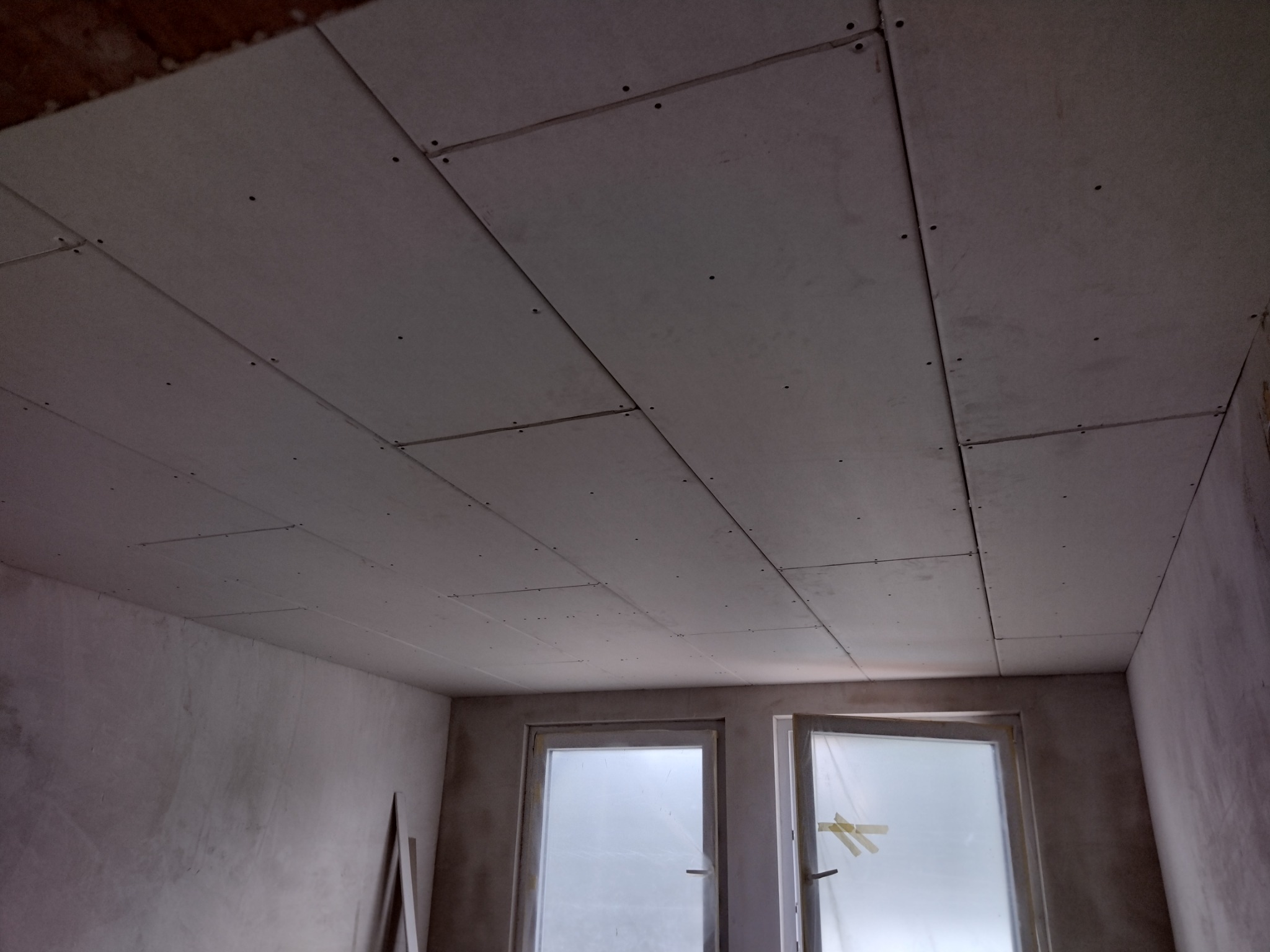 Płyty kartonowo-gipsowe zamontowane na suficie, widoczne łączenia i ślady po wkrętach, ściany w stanie surowym, okna zabezpieczone folią.