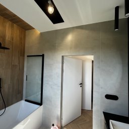 Marcin Suchański - Nowoczesna łazienka z wanną z czarną kabiną, szarymi płytkami, drewnopodobnymi panelami, oświetleniem punktowym i otwartymi drzwiami do kolejnego pomieszczenia z parkietem w jodełkę.