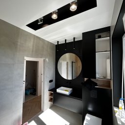 Marcin Suchański - Nowoczesna łazienka w szaro-czarnej kolorystyce z okrągłym lustrem, punktowym oświetleniem i widoczną chemią budowlaną w rogu, podkreślająca etap wykończenia.