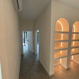 Marcin Suchański - Jasne wnętrze z szarymi płytkami podłogowymi, widoczne drzwi balkonowe w głębi i podświetlane półki w kształcie łuków po prawej stronie, klimatyzator na ścianie po lewej.