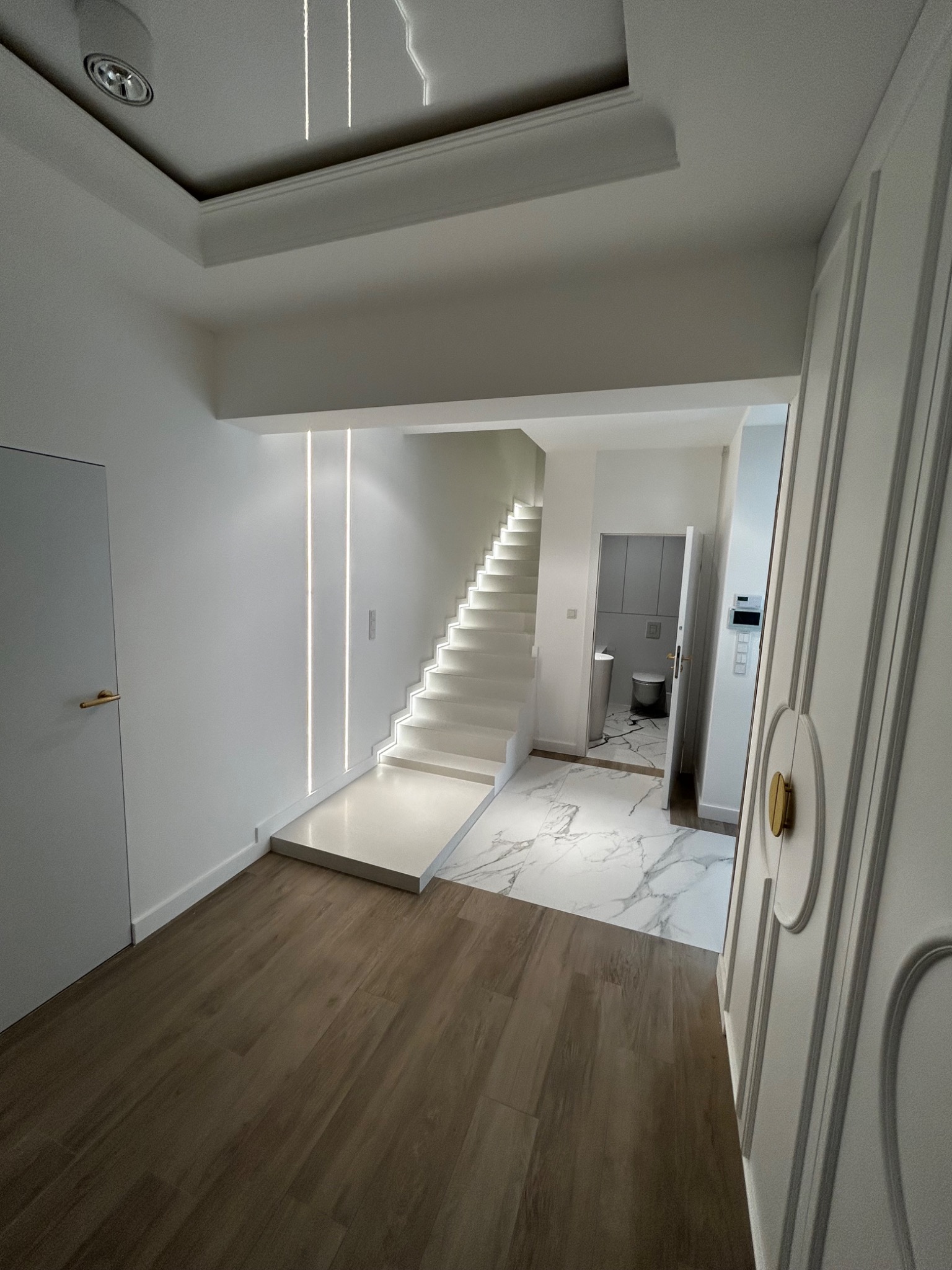 Nowoczesne wnętrze: białe schody z podświetleniem LED, marmurowa podłoga przechodząca w drewnopodobne panele, widok na minimalistyczną łazienkę z toaletą i szafkami.