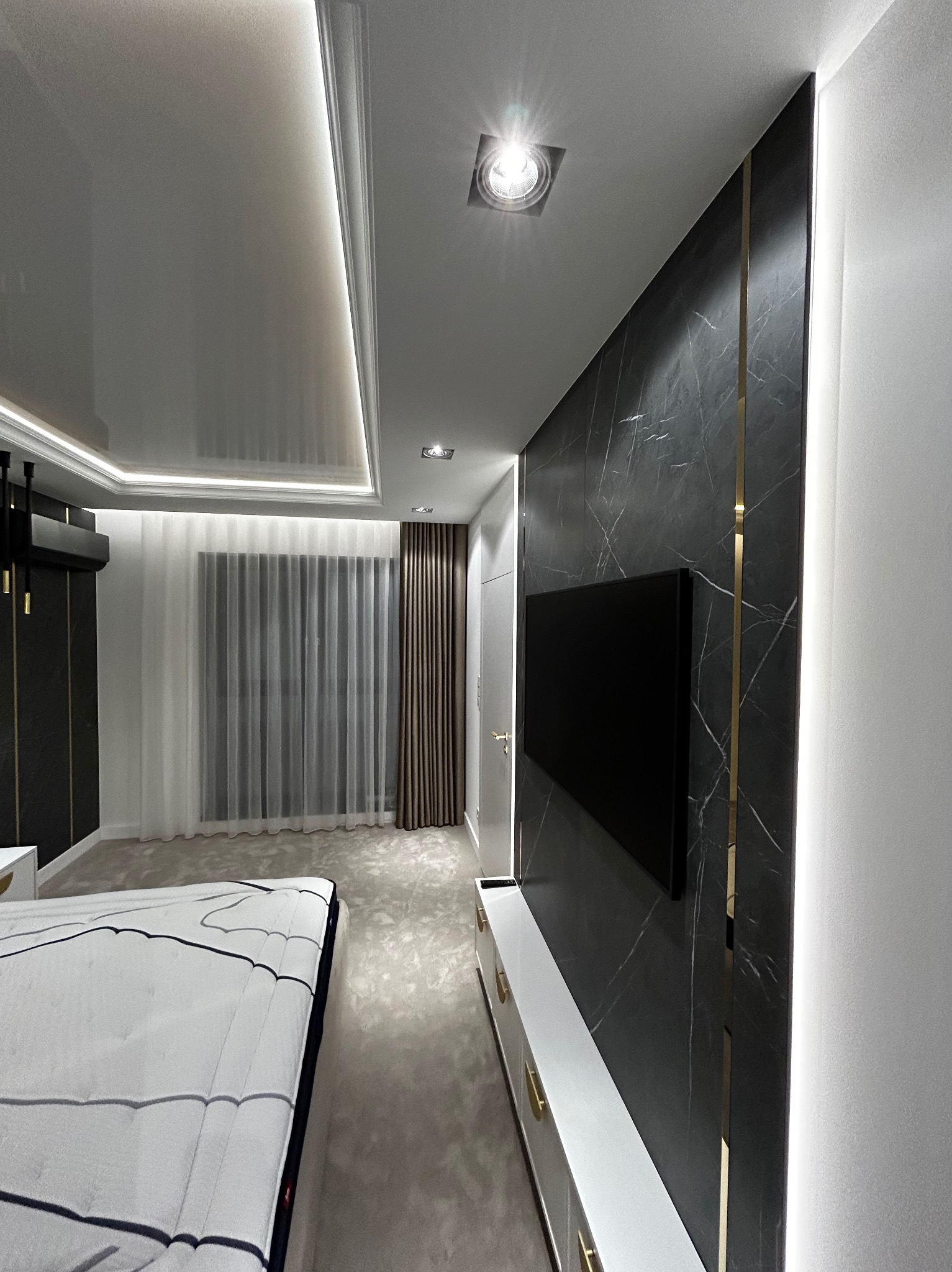 Nowoczesna sypialnia z czarną ścianą z marmuru, telewizorem, białymi meblami i podświetlanym sufitem oraz firanami.