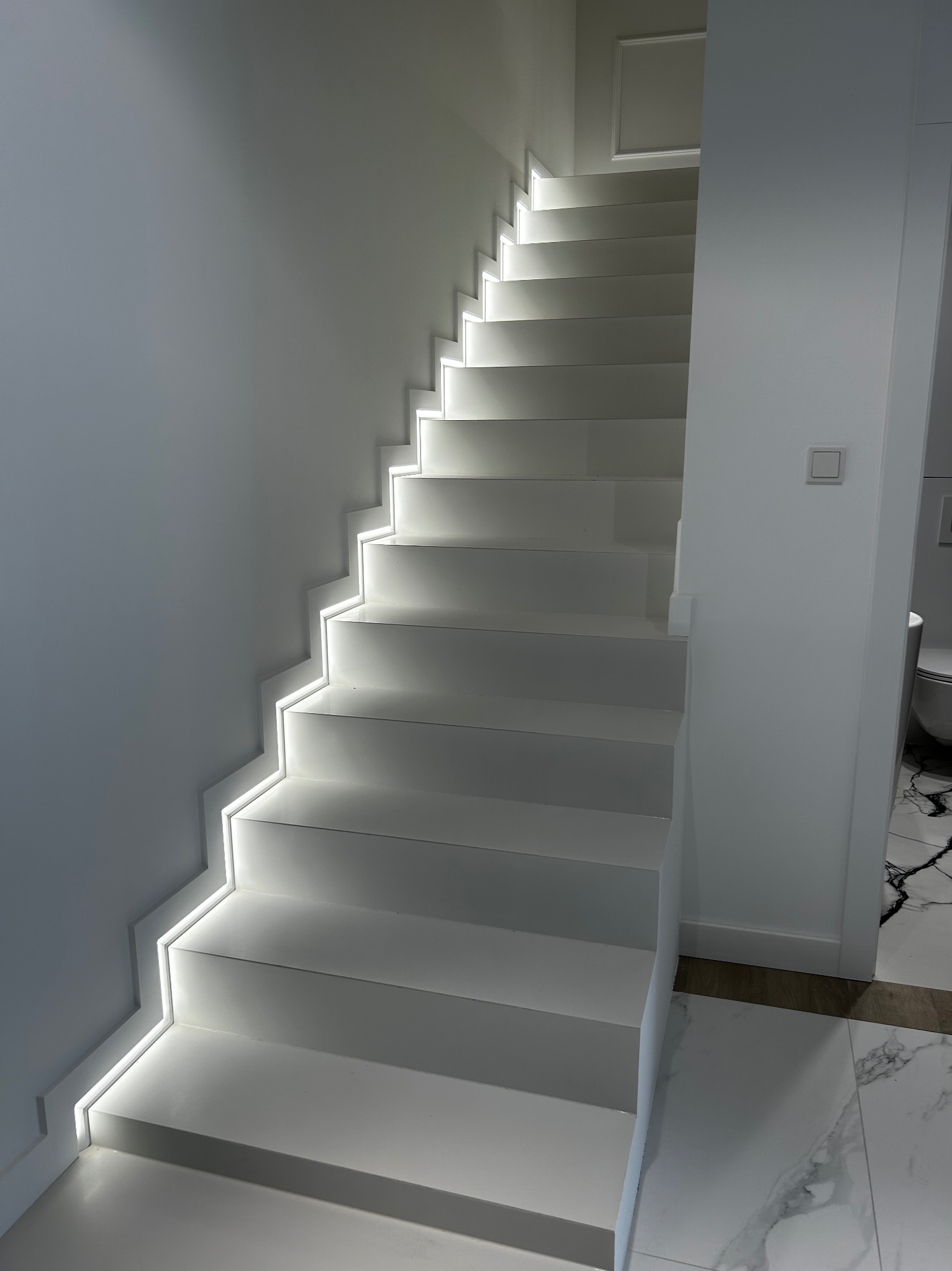 Nowoczesne, białe schody z subtelnym oświetleniem LED wzdłuż stopni, prowadzące w górę, z fragmentem łazienki z marmurową podłogą widocznym w tle.