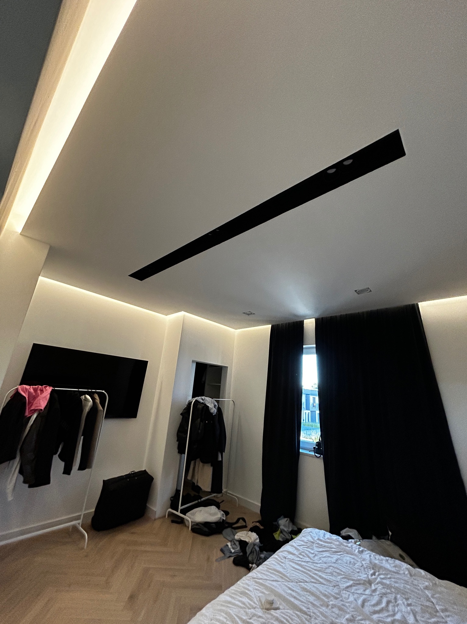 Nowoczesna sypialnia z podświetleniem LED wzdłuż sufitu i ścian, czarnymi zasłonami i drewnianą podłogą w jodełkę; widoczne ubrania na wieszakach i minimalistyczny wystrój.