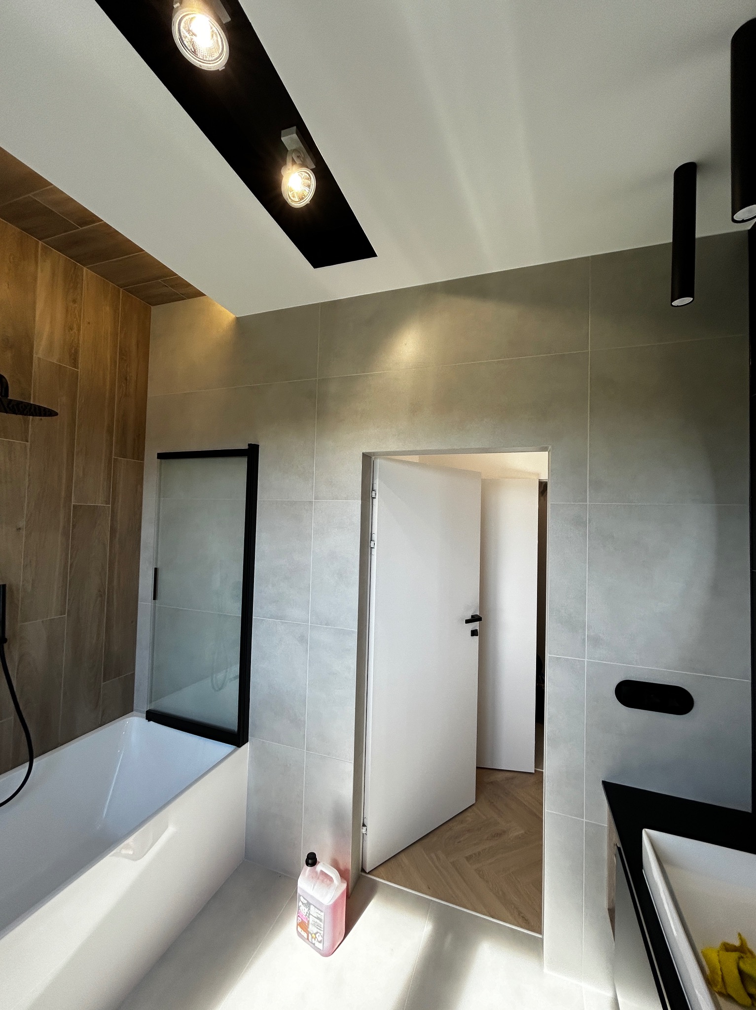 Nowoczesna łazienka z wanną z czarną kabiną, szarymi płytkami, drewnopodobnymi panelami, oświetleniem punktowym i otwartymi drzwiami do kolejnego pomieszczenia z parkietem w jodełkę.