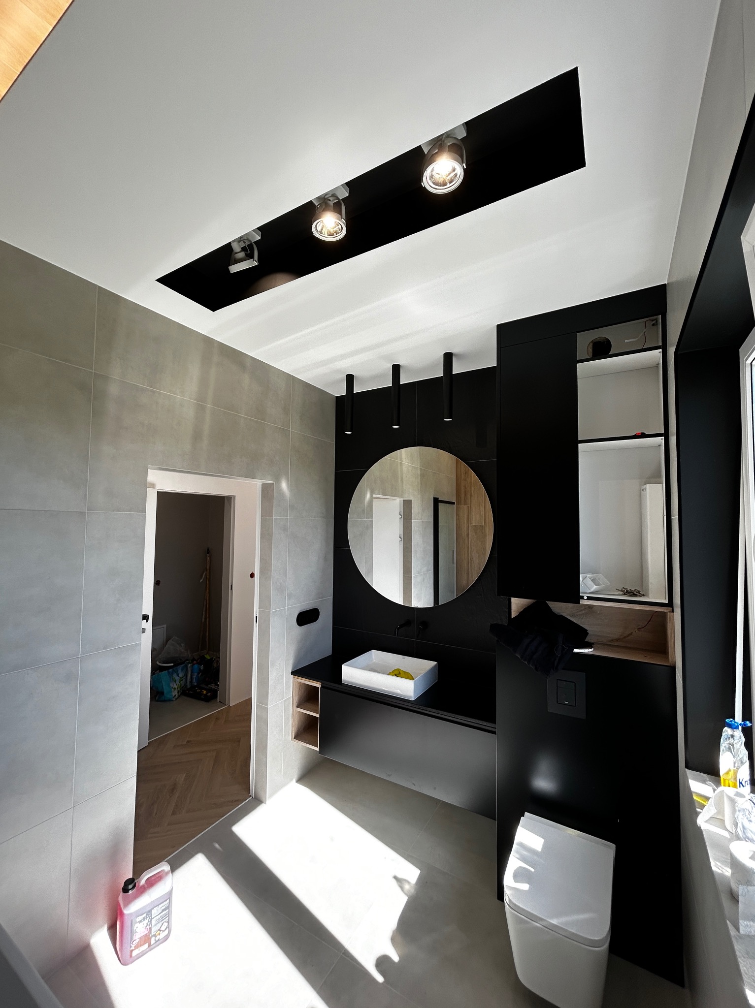 Nowoczesna łazienka w szaro-czarnej kolorystyce z okrągłym lustrem, punktowym oświetleniem i widoczną chemią budowlaną w rogu, podkreślająca etap wykończenia.