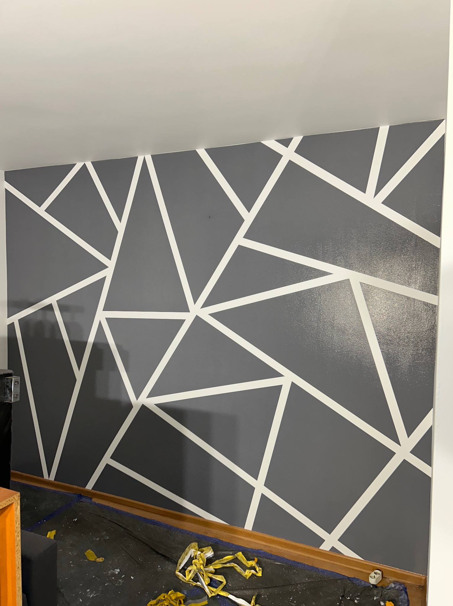 Ściana pomalowana na szaro z białymi geometrycznymi wzorami, zabezpieczona folią malarską na podłodze, widoczne pozostałości taśmy malarskiej.