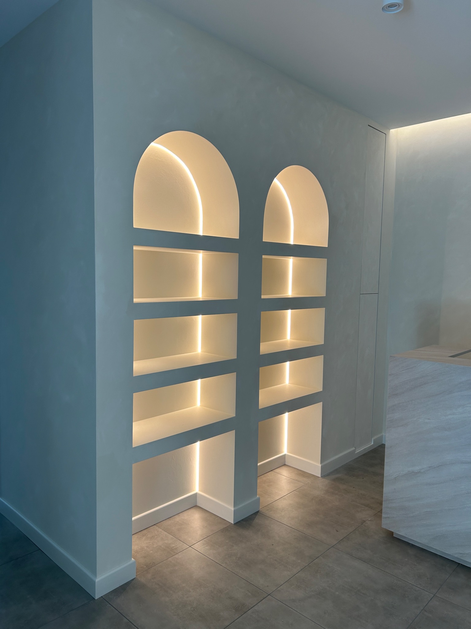 Dwa podświetlane, wnękowe regały z półkami w kształcie łuków w nowoczesnym wnętrzu, oświetlenie LED podkreśla minimalistyczny design i szarą kolorystykę ścian oraz podłogi.