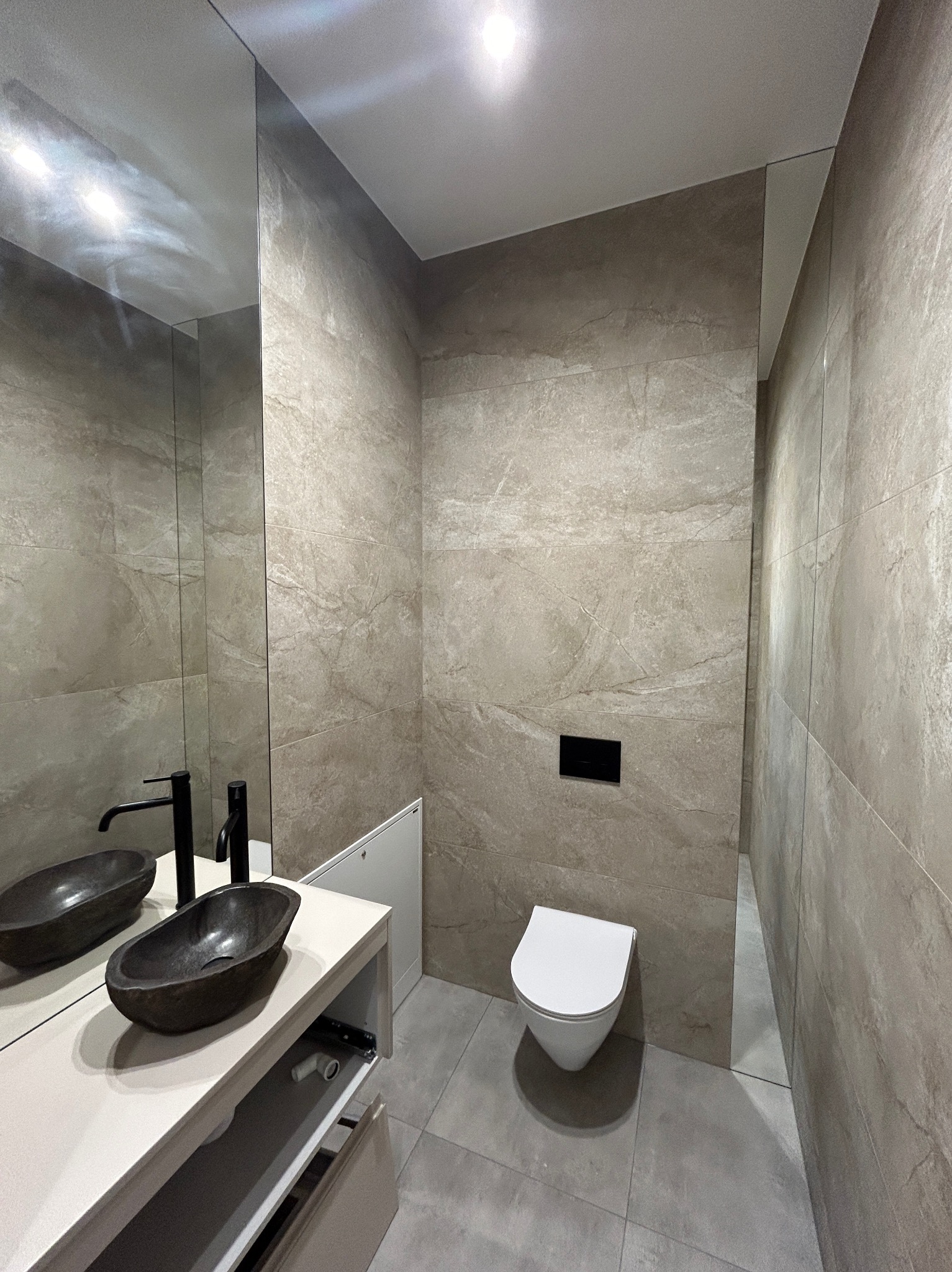 Nowoczesna łazienka z kamienną umywalką w kształcie łzy, czarną armaturą, płytkami imitującymi kamień na ścianach i podłodze, oraz podwieszaną toaletą z czarnym panelem sterującym.