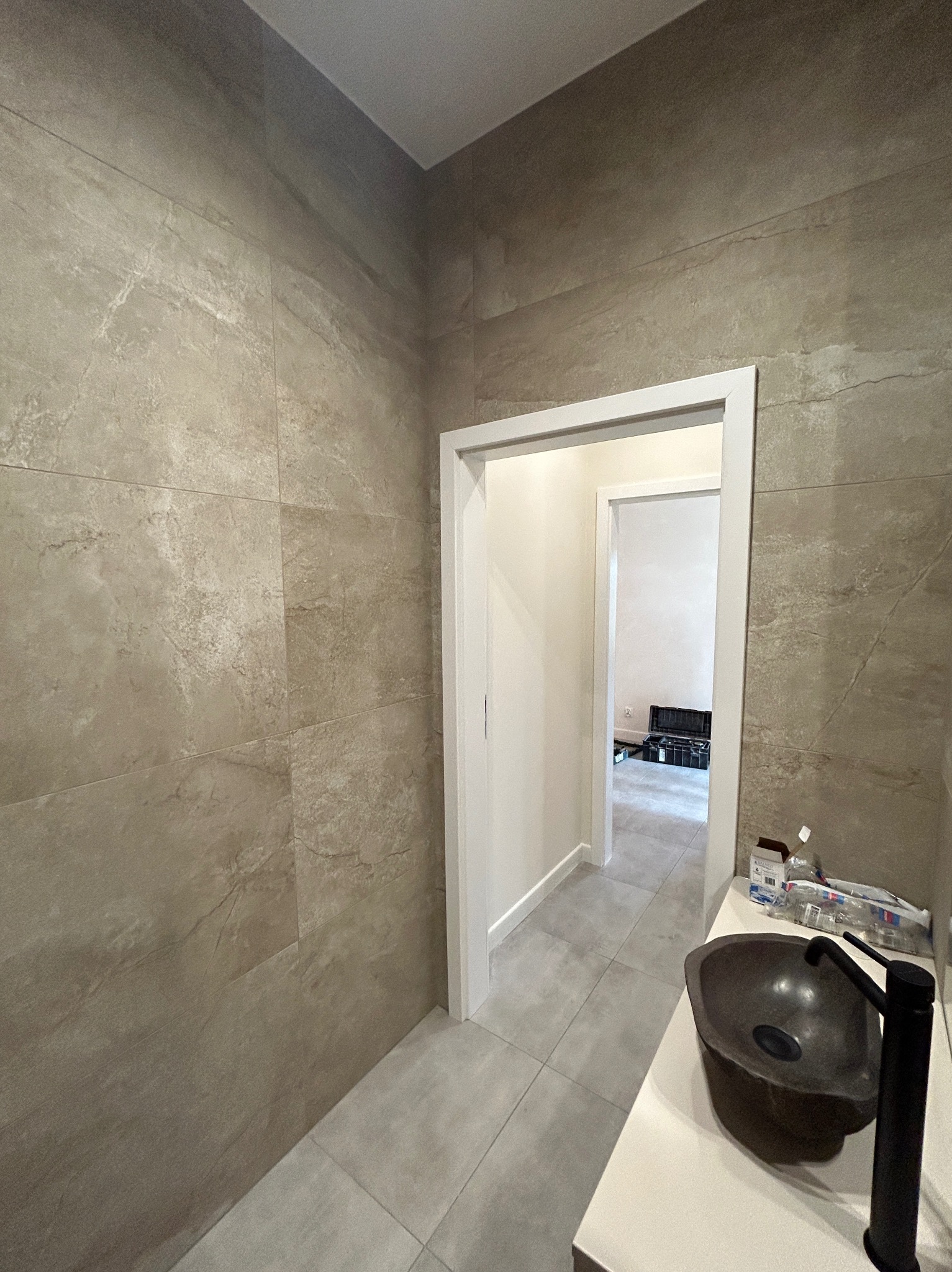 Wykończona łazienka z kamienną umywalką na blacie, szare płytki na ścianach i podłodze, widok na korytarz przez białe futryny drzwiowe.