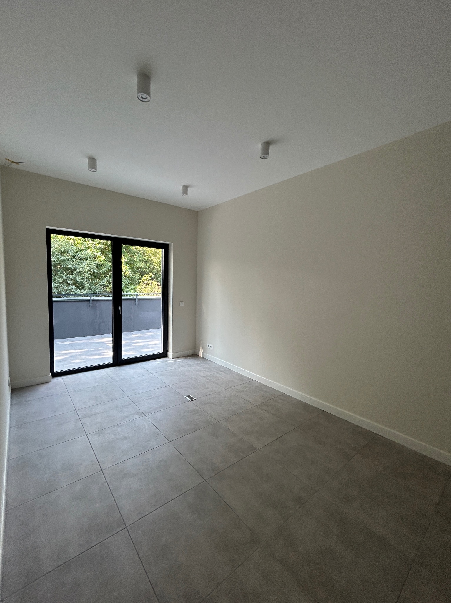 Pusty, jasny pokój z szarymi płytkami podłogowymi, białymi ścianami i dużym, czarnym oknem balkonowym z widokiem na zieleń. Na suficie widoczne okrągłe lampy punktowe.