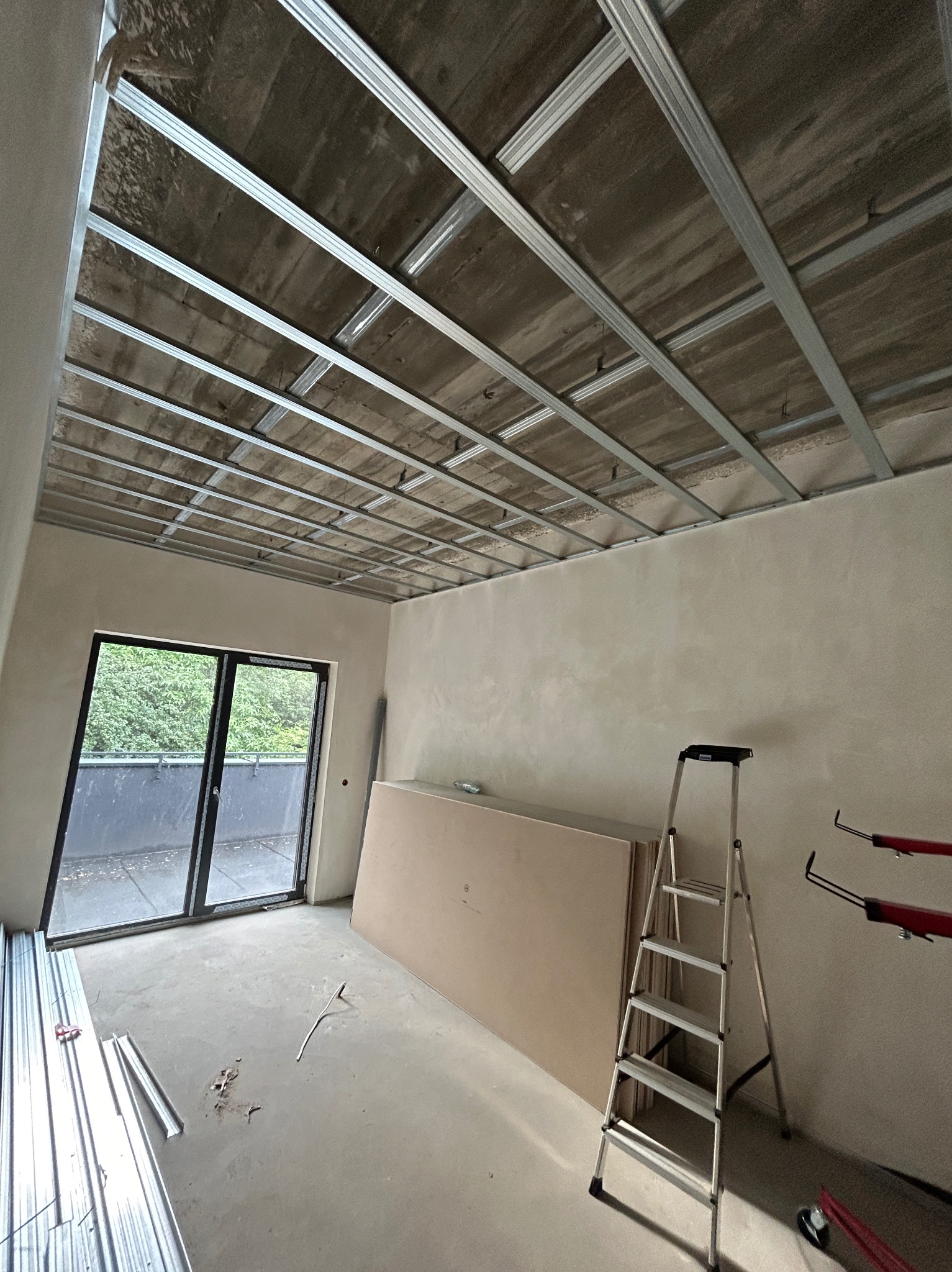 Wnętrze w trakcie prac wykończeniowych: metalowa konstrukcja sufitu podwieszanego, płyty kartonowo-gipsowe oparte o ścianę, aluminiowa drabina rozstawiona w pomieszczeniu z dużym oknem balkonowym.