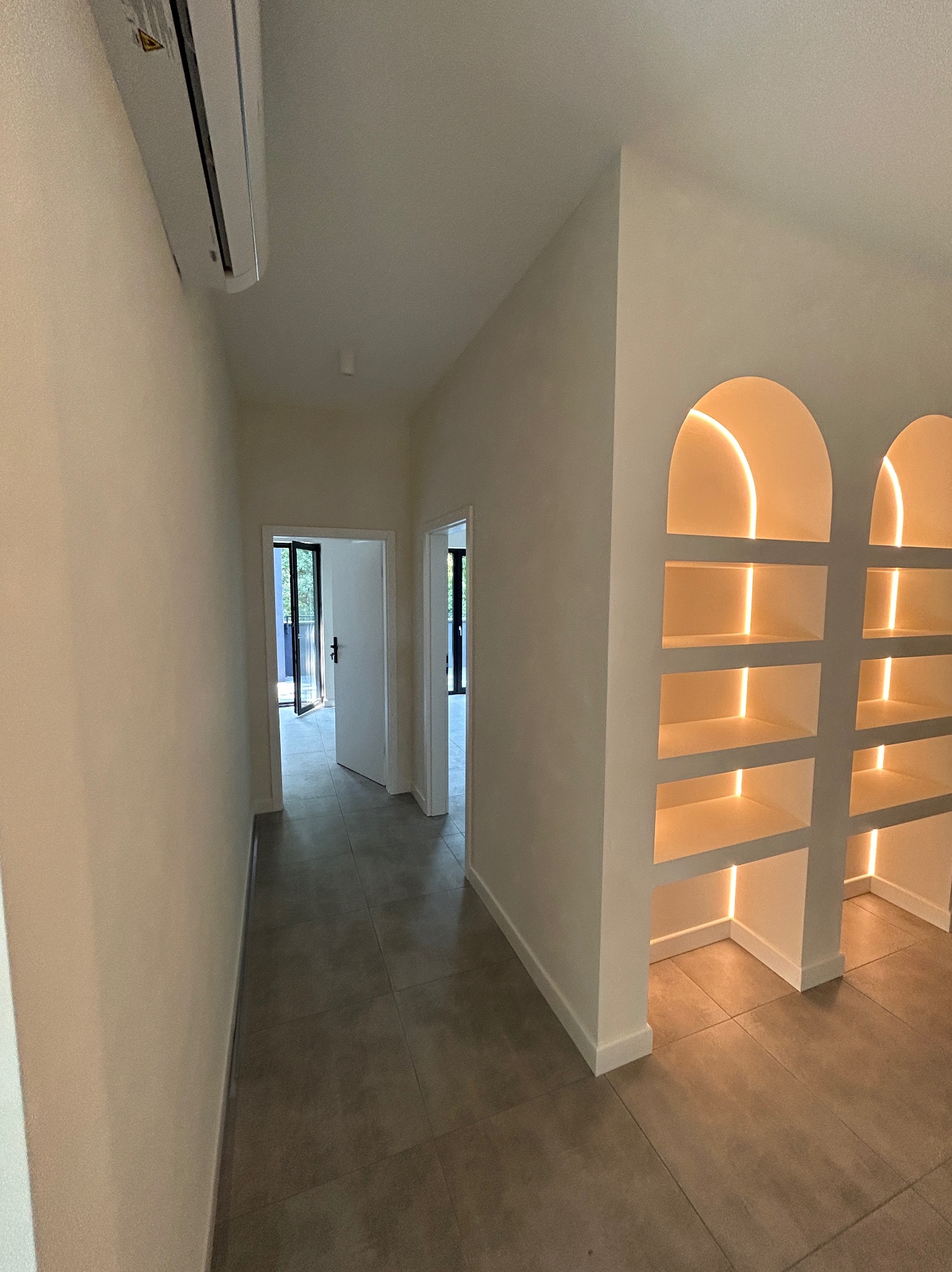 Jasne wnętrze z szarymi płytkami podłogowymi, widoczne drzwi balkonowe w głębi i podświetlane półki w kształcie łuków po prawej stronie, klimatyzator na ścianie po lewej.